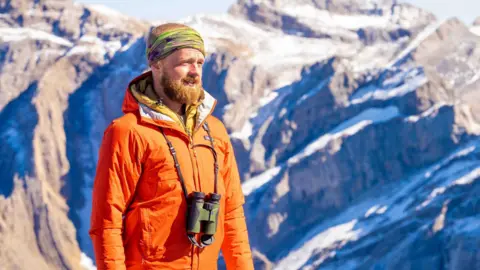Will Hawkes Photography Naturalist Will Hawkes on a hike. He stands in front of rocky snow-dusted mountains, wearing an orange coat and a green bandana on his head. He has a pair of binoculars around his neck.