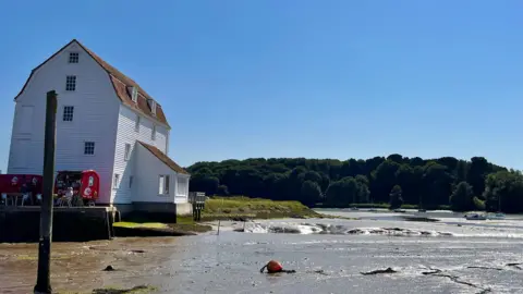 On the left is a large white mill building.  It's on the edge of the river and is around four or five storeys high. It's white and is made up of horizontal wooden slats.  There are four small windows on the side of the building and a further five on the front with two windows in the tiled roof.  The tide is out and the mud of the river bed is visible. There are a lot of trees in the background and a blue, cloudless sky.