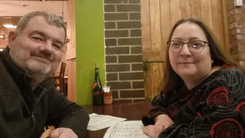Andrew Palmer A man with grey hair and facial hair sitting across the table from a woman with brown hair and glasses. They are taking a selfie and there are white menus on the table in front of them.