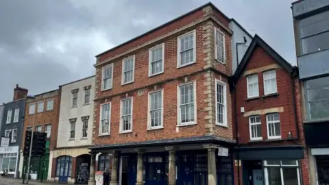 1625 Independent People Modern day Kingsley Hall as seen from the outside. The upper two storeys are made of red brick, with large rectangular white Georgian windows. The top two storeys jut out over the pavement below, resting on five columns. The ground floor, which is in shadow, is set back, in line with other town house buildings on the street.