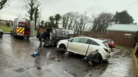 A number of cars in various colours are parked in a car park. A work van is seen in the background as workers attempt to fix tyres. 