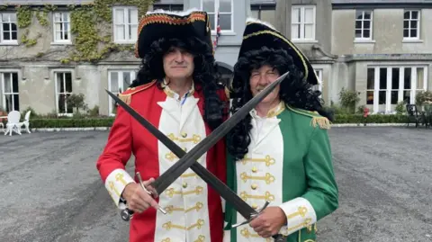 Two actors dressed as King William III and James II. William is dressed in red and white and James in green and white. Both are wearing long black wigs and are crossing swords. 