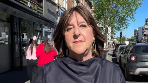 Judith Nemeth is pictured looking directly at the camera. She is standing in Golders Green and is wearing a draped black top, gold dangly earrings and has short brown hair with a fringe.