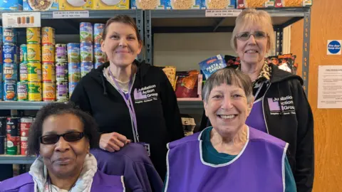 Four women photographed in front of a shelving unit stacked with food items, mostly tins. They are wearing purple tabards with "Jubilation Action Group" logos on them.