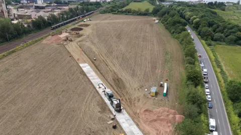 Network Rail Aerial view of a large brown field with a straight temporary concrete roadway running diagonally across it. It connects a road and the railway on either side of the field.
