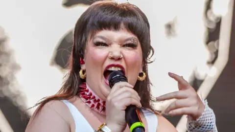 Getty Images Lola Young singing on stage with her mouth open, one hand holding a microphone and the other gesturing