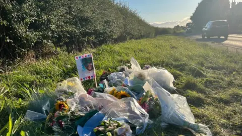 In the foreground are about 7 or 8 bunches of flowers laid on a grassy verge. There's an image of Wayne Neville on a white background with pink and blue hearts around the edge. The A53 is just visible in the top right corner, with a black car driving along it. The sky is blue with some clouds, with low sunlight suggesting it's late in the day. 