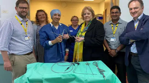 Male and female surgeons and volunteers standing behind a table covered with a green cloth and medical equipment. They are smiling.