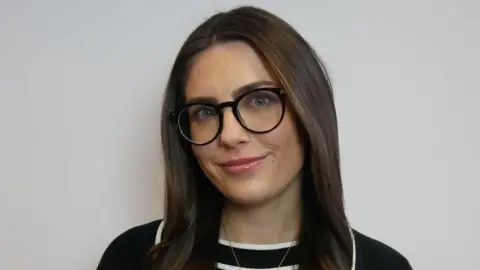 VerriBerri Amy Spooner, who has long brown hair and is wearing glasses with black rims. She is also wearing a black top with white stripes through the middle. She is standing in front of a grey wall.