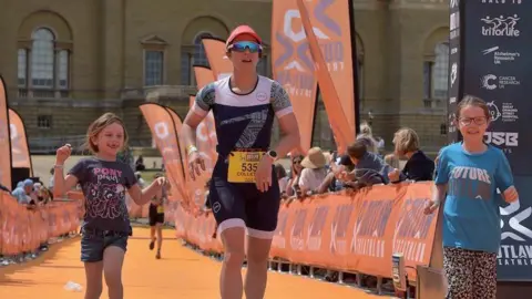 Mike Green Collette runs over the finish line of a triathlon event with her two daughters. The daughters are smiling as they run with her.
