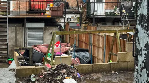 The back of some premises which have railings and metal steps. At the bottom of one are children's swings. In front of these is a pile of rubbish which includes furniture. In front of the swings, near to the camera, is a smaller pile of dirt and rubbish and some puddles and mud. 