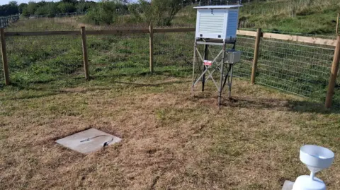Crown Copyright Inside a small boundary, fenced off with wood and chicken wire, there is a wooden box propped on stilts, a concrete square on the ground, and a small white rain gauge