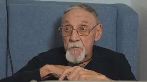 Alex Smith looking slightly away from the camera. He has glasses on and short grey hair. He also has a white beard. He is wearing a dark top and is sitting on a blue chair. His hands are folded in front of his body.