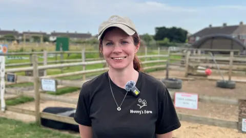 A woman wearing a neutral-toned cap and brown hair smiles to the camera. She is wearing a black T-shirt with the words 'Nunny's Farm' printed in white text on her chest.
She is stood on the farm where pig pens are in the background amongst other enclosures.