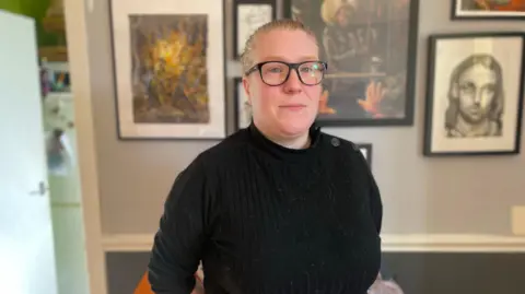 Kirsty Pattison standing in front of a wall with multiple pictures. It is two tones of blue separated by a white wooden line. Kirsty has fair hair pulled back. She is wearing dark-rimmed glasses and a black top with black buttons on the right shoulder.