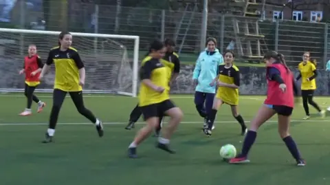 BBC TV screenshot from the edge of a football pitch on which a training session is taking place. Female players wearing red bibs or yellow football tops are kicking the ball.