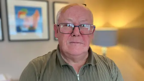 Ted Milward is wearing square glasses and a green sweatshirt. He is in his living room and there are pictures and a lamp in the background. 