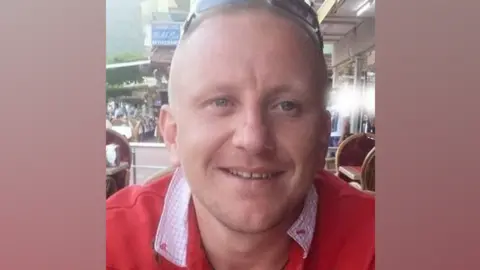 A man with cropped fair hair and wearing a red jumper and sunglasses on his head smiles at the camera. He is sitting on a restaurant terrace. 