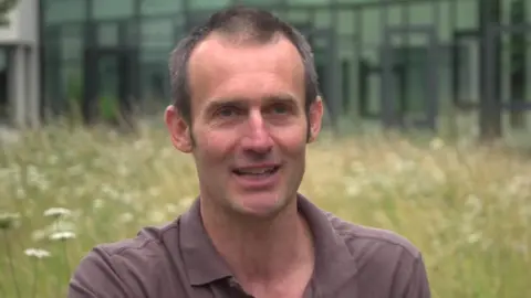 A head-and-shoulders photo of a man with short dark greying hair and wearing a light brown polo shirt, with a flowery stretch of grassland and a building in soft focus behind him.