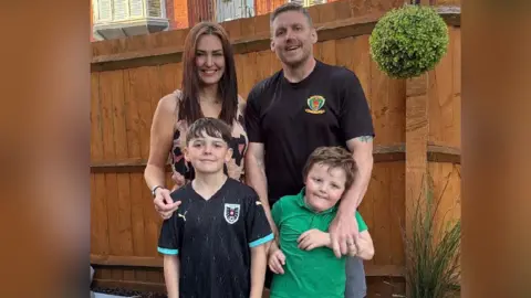 Robert Cummings Family picture of Ellis with his brother, mum and dad. He is standing on the left next to his brother, with Gemma and Robert standing behind them. They are standing in front of a brown fence in a garden. They are smiling and looking at the camera.