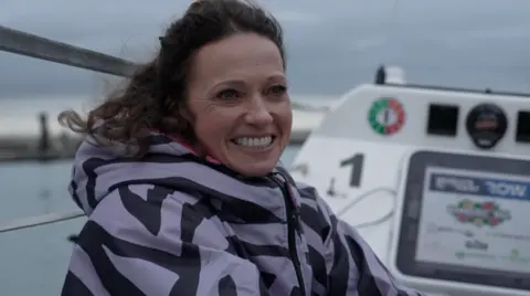 A woman with brown hair smiling at the camera and sat in a row boat.