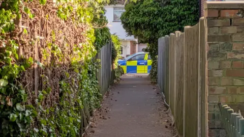 Hilary Silvester A police car at the end of a path, with fences either side, and trees and bushes either side.