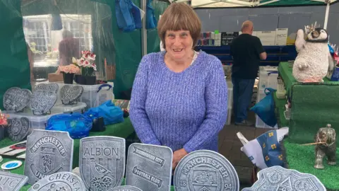 BBC A woman standing in a market stall - in front of her are a series of ornaments of football badges, including Manchester United, West Bromwich Albion and Birmingham City. She is wearing a purple jumper, and behind her is another trader with his back turned.
