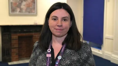 John Fairhall/BBC A woman with shoulder-length black hair. She has a purple lanyard around her neck. She is looking into the camera. 