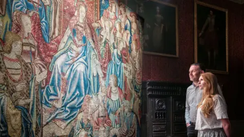 A man and woman are looking at a Tudor tapestry which depicts a scene from the Old Testament Book of Esther and is blue and red in colour.