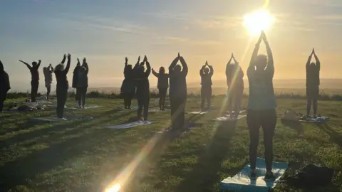 Beachy Head Ambassadors We see around 15 people doing a yoga class. We see them from behind with their hands above their heads and their palms together in a prayer pose. They are on the clifftop at Beachy Head and they are silhouetted against the sun setting out to sea on the horizon.