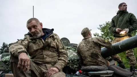 Getty Images Ukrainian soldiers in full battle dress sit on a cannon in a muddy field.  