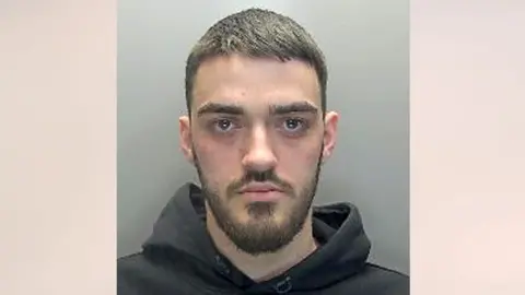 Lirim Shiqerukaj has short brown hair, and a moustache and beard. He is wearing a black hoodie and looking straight at the camera without any expression.