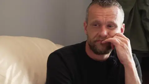 A bearded man looks down with his hand to his mouth.