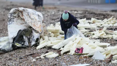Eddie Mitchell A woman in a black coat and green woolly hat is clearing up large foam washed up on a pebbled beach