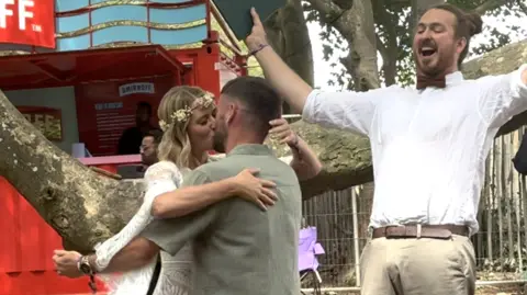 Craig Brown, who has brown cropped hair and is wearing a khaki green shirt, and Tara Sweeting, who has long blonde hair and is wearing a white lace dress, embrace and kiss in front of a man, wearing a bow tie. He is cheering and has his arms aloft.