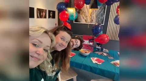The Helm Two women and a man peer into the frame from the side, with a table at the centre of the image surrounded by red and blue balloons and leaflets advertising The Helm.