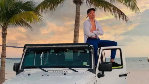 instagram Samy Daim sitting on top of a white 4x4 vehicle parked beside a beach with palm trees in the background. Mr Daim is wearing an unbuttoned white shirt with blue trousers and cap.