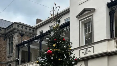 The top of a decorated Christmas tree on Market Street in St Peter Port, Guernsey. There is gold tinsel wrapped around with red and white baubles. There is a gold star at the top and buildings in the background 