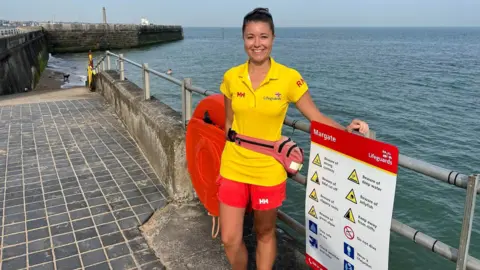 BBC Kent A RNLI lifeguard stands by metal railings with the sea behind her.