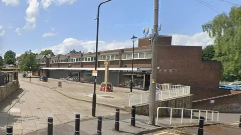 An empty suburban parade of shops. There is a war memorial, lampposts and other street furniture.