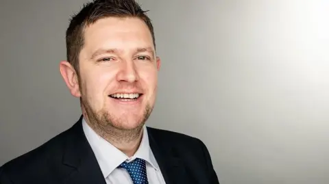 NIS Group A man with dark hair, swept to his right side and with dark stubble, smiles at the camera. He wears a dark suit, white shirt and blue tie with white dots on it. Behind him is a grey wall.