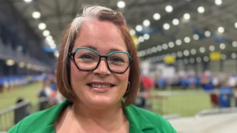 Sian is smiling at the camera at a large centre blurred out in the background with lights on the roof.
She is wearing an emerald green blazer, blue and purple rounded glasses and her hair is face length with dark orange reddish hair with white highlights at the fringe and front layer.