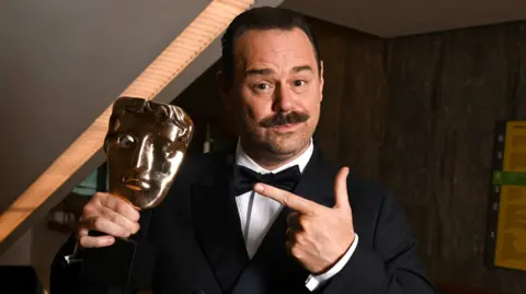 Getty Images Danny Dyer in a black tux points to his Bafta which he is holding in his left hand. He has a moustache.