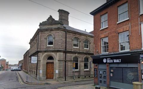 Macclesfield library on the corner of a street in an old bank building with a brown wooden door. It is situated opposite the Beer Vault bar. 
