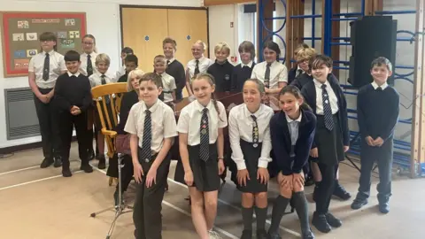 BBC A group of schoolchildren stand around a piano in a school sports hall . Singer Gwenno sits at the keyboard with a microphone