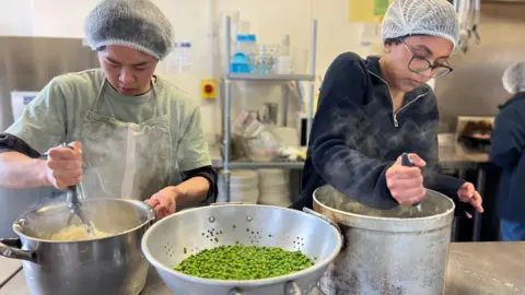 Two students wearing white hair nets stirring pots with hot food. There us a colander full of green peas in front of them. Steam is rising from their pots. Someone else could be seen behind them.