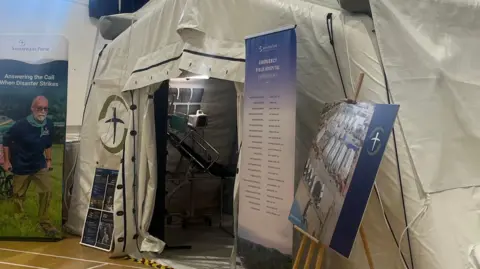 A large white tent structure is in a sports hall.
A number of signs and billboards are seen near the entrance.
The front door of the tent is open and a makeshift hospital bed and medical instruments are visible.