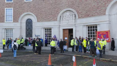 LDRS Around 30people holding banners and flags with union names and logos, some wearing hi-visibility jackets, standing outside county hall in Taunton