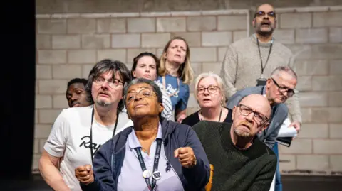 Graeme Braidwood Photography A group of actors in a group together. The woman in front looks like she is holding a steering wheel. Their faces look surprised and are mainly looking to the left. 