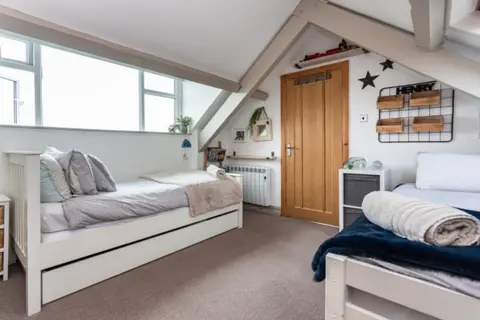 Michael Speak A bedroom with a slanted ceiling and two single beds, one on each side of the room. In front is a wooden door and a chest of drawers, and pictures and storage hanging on the wall. 
